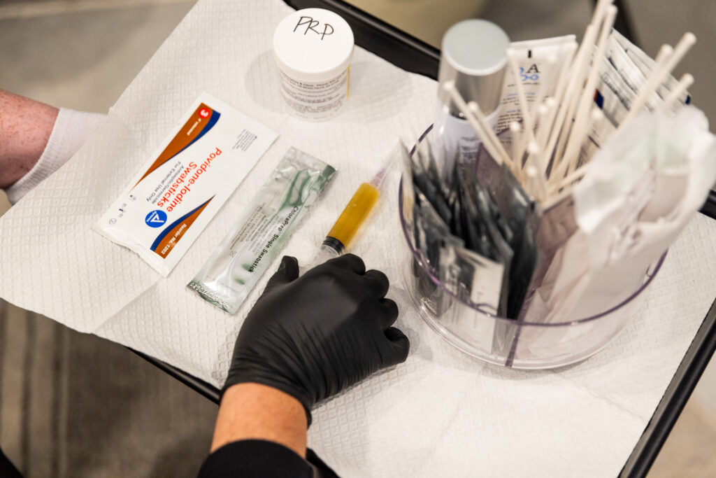 A Mayo stand, holding all the tools necessary for PRP shots in Sioux Falls, including a prepared PRP shot, disinfecting wipes, and cotton swabs. The hand of a provider, wearing a surigical glove, is reaching for the PRP shot to prepare for the treatment. 